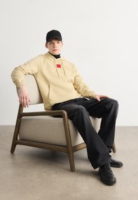 Sudadera beige con un bolsillo frontal y un parche con logo rojo, combinada con pantalones negros y zapatos negros, sentado en una silla clara con un marco de madera.