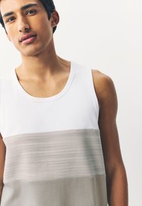 Bezroku tops ar baltu augšdaļu un virs tās horizontālu teksturētu bēšas krāsas svītru, izgatavots no gludas kokvilnas auduma.