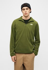 Νέος άνδρα ς που φοράει μαύρο καπέλο, φλις σε ελιά πράσινο με φερμουάρ μέχρι το στήθος και το λογότυπο The North Face, και μπεζ παντελόνι, στέκεται με το ένα χέρι στην τσέπη.