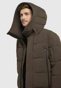 Smeđi pufasti kaput s velikim, kockastim kapuljačom. Ima džepove sa zatvaračem i pun zatvarač sprijeda, izrađen od teksturiranog materijala.