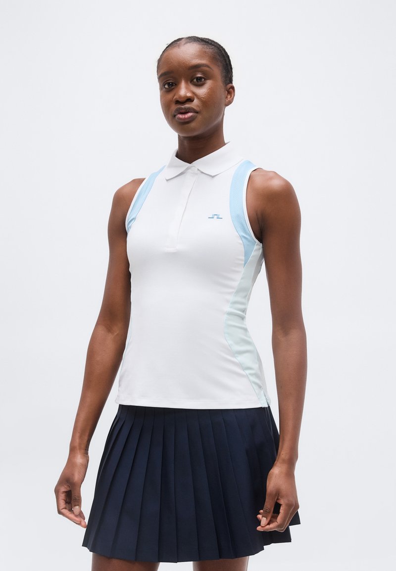 Νεαρή γυναίκα που φοράει λευκό αμάνικο πόλο με ανοιχτό μπλε πάνελ και μπλε σκουρόχρωμη πλισέ φούστα, στέκεται μπροστά σε απλό φόντο.