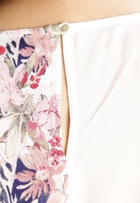 Primer plano del escote trasero de una prenda que muestra un pequeño botón que sujeta una abertura entre paneles de tela blanca con flores y lisa.