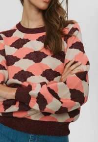 Ženska z dolgimi rjavimi lasmi, oblečena v pulover z vzorcem v roza, beži in temno rjavi barvi, s prekrižanimi rokami na enobarvni osbackground.