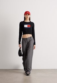 Černý zkrácený svetr s logem "TOMMY JEANS", šedé pruhované široké kalhoty, červená čepice a černá kabelka. Model má na sobě černé baleríny.