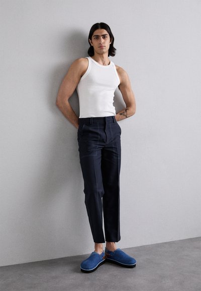 White ribbed tank top, navy tailored trousers with contrasting stitching, and blue slip-on shoes with brown accents against a gray background.