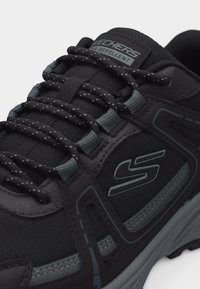 Μαύρο αθλητικό παπούτσι Skechers με γκρι λεπτομέρειες, αντανακλαστικά κορδόνια με τελείες, ετικέτα «Απωθητικό Νερού» και εμφανές υφή σόλας σε λευκό φόντο.