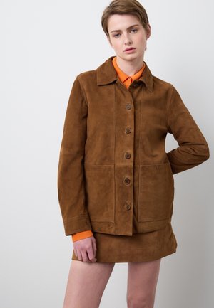 Braune Wildlederjacke mit Knopfleiste, spitzem Kragen und zwei Vorderentaschen. Trägt man über einem leuchtend orangefarbenen Hemd. Glatte Textur.