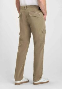 Man som bär beige cargobyxor med ficklock, vita sneakers och en vit skjorta, stående mot en enfärgad ljus bakgrund.