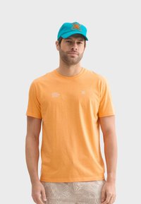 Camiseta de algodón naranja con mangas cortas y cuello redondo, que presenta texto blanco en la parte delantera. Se lleva con una gorra azul que muestra un logo decorativo.