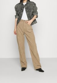 Giacca di denim nera con chiusura frontale a bottoni, due tasche sul petto; abbinata a pantaloni largi in velluto a coste marrone chiaro. T-shirt bianca tinta unita indossata sotto.