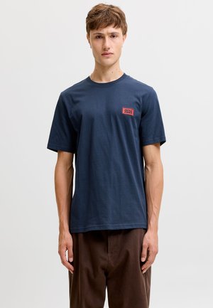 Jeune homme debout, face à l'avant, portant un t-shirt bleu foncé à manches courtes avec un petit logo rouge "Jack & Jones" sur la poitrine et un pantalon marron.