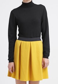 Schwarzer Rollkragenpullover kombiniert mit einem gelben Plisseerock. Der Rock hat einen schwarzen Taillenbund und einen strukturierten Stoff.