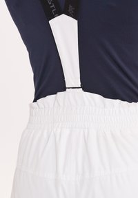Witte overalls met elastische tailleband en verstelbare witte bretels tegen een marineblauw shirt, met gladde stof en een minimalistisch ontwerp.