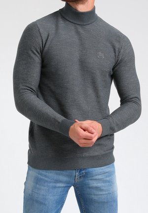 Pull en col roulé gris, à côtes, avec un motif texturé, une coupe ajustée et un petit patch logo sur la poitrine. Associé à un jean bleu.
