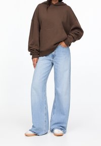 Sweat à capuche marron avec épaules tombantes, associ é à un jean large bleu clair. Le tissu semble doux, avec une coupe décontractée.