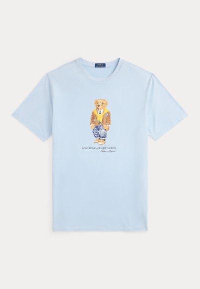 Hellblaues kurzärmeliges T-Shirt mit einem in der Mitte aufgedruckten Polo-Bär, der eine braune Jacke, einen gelben Schal, blaue Jeans und hellbraune Schuhe trägt.