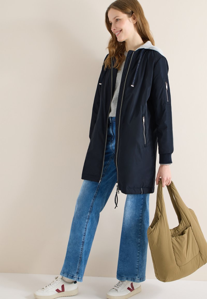 Jeune femme portant un manteau à capuche bleu marine, un sweat à capuche gris clair, un jean large bleu, des baskets blanches, tenant un grand sac cabas beige matelassé, souriante et debout.