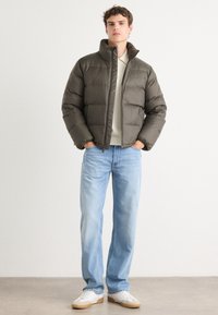 Veste matelassée vert foncé avec un col haut, fermeture éclair et poignets élastiques, portée sur un pull léger et associée à un jean bleu et des baskets blanches.