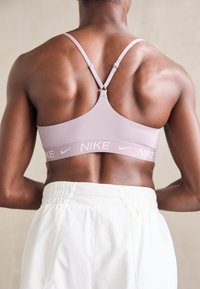 Sujetador deportivo lila claro con tirantes ajustables y una banda elástica con marca. Combinado con pantalones blancos de cintura elástica, que muestran un tejido texturizado.