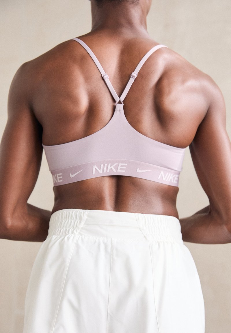 Sujetador deportivo lila claro con tirantes ajustables y una banda elástica con marca. Combinado con pantalones blancos de cintura elástica, que muestran un tejido texturizado.