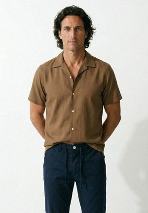 Hombre con cabello oscuro que lleva una camisa marrón de manga corta abotonada y pantalones azul marino, de pie con las manos detrás de la espalda sobre un fondo blanco.