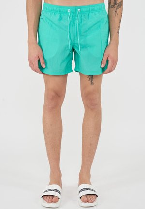 Man draagt turquoise zwembroek met witte trekkoorden en witte Emporio Armani slides, staand tegen een effen witte achtergrond.