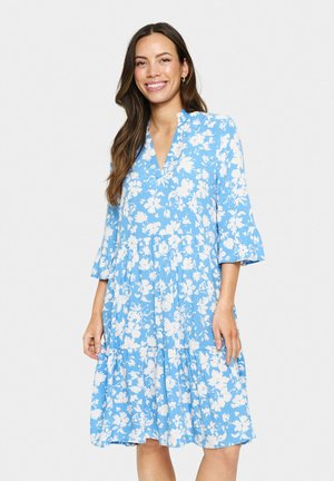 Blå blommig klänning med V-urringning, trekvarts armar med klockform, lager på lager-skirt och vita blommönster över en strukturerad tyg.