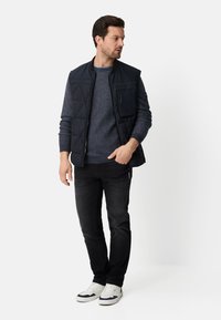 Granatowa pikowana kamizelka z kieszenią na zamek, noszona na ciemnoniebieskim swetrze z dzianiny, w połączeniu z czarnymi dżinsami i białymi sneakersami.