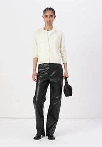 Gilet créme avec des boutons, associé à un pantalon en cuir noir. Le mannequin tient un sac noir texturé, portant des bottines noires. Fond uni.
