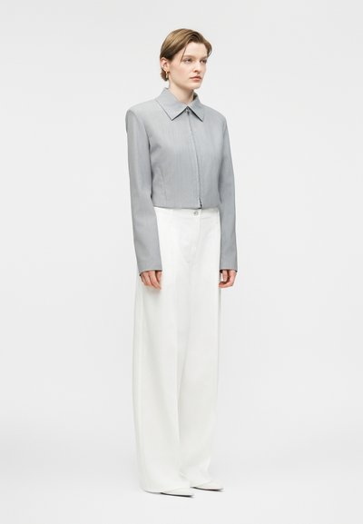 Γυναίκα που φορά γκρι κοντό μπουφάν με μακριά μανίκια και λευκό παντελόνι με φαρδιά μπατζάκια, μπροστά σε λευκό απλό φόντο.