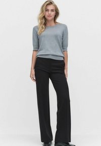 Kvinna stående iklädd en grå kortärmad tröja, svarta vida byxor och svarta skor mot en ljust enfärgad bakgrund.