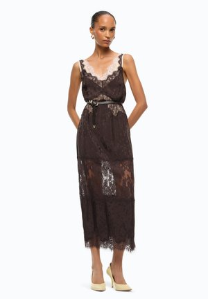 Femme portant une robe noire en dentelle sans manches avec des panneaux transparents, une ceinture noire et des escarpins beiges, debout devant un fond blanc.