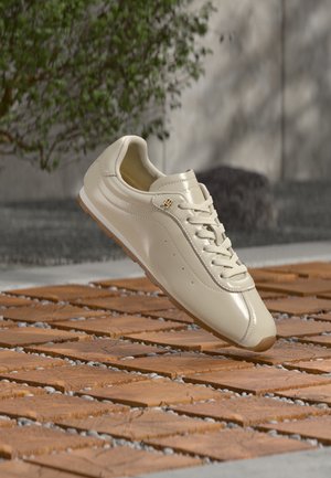 Beige lederen sneaker met veters en een klein goudkleurig logo, zwevend boven een patroon van hout en grind buiten.