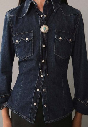 Kvinde iført mørkeblå denimskjorte med trykknapper, to brystlommer og et bolo-slip med turkis sten ved kraven.