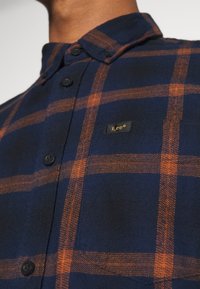 Gros plan d'une personne portant une chemise à carreaux bleu marine et orange avec une petite étiquette noire portant la mention "Lee" sur la poitrine.