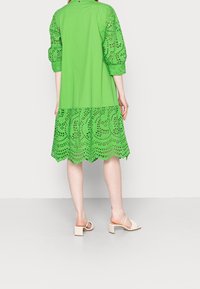 Mujer vistiendo un vestido verde brillante hasta la rodilla con mangas y dobladillo de encaje, combinado con sandalias de tacón beige, de pie sobre un suelo blanco.