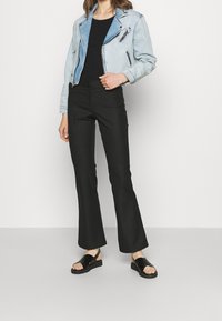 Světlomodrá denimová motorkářská bunda s detaily na zip, zkombinovaná s černými rozšířenými kalhotami a černými plochými sandály. Hladké textury, volný střih.