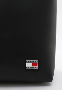 Schwarze synthetische Tasche mit glatter Textur. Verfügt über ein silbernes Tommy Jeans Logo-Patch mit roten und weißen Akzenten. Kompaktes Design.