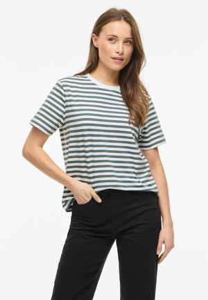 Femme portant une chemise à manches courtes blanche et verte à rayures avec un pantalon noir, debout avec une main dans la poche devant un fond uni.