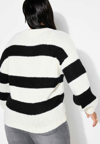 Maglione a righe bianche e nere in maglia, con vestibilità ampia, polsini e orlo a coste. Texture visibile dal filato spesso, indossato sopra jeans grigi.