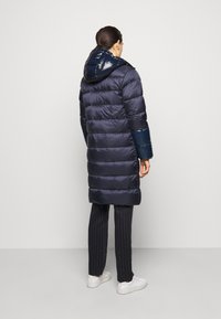 Marinblå puffjacka med glänsande huva och ärmar, horisontellt kviltad design och lång silhuett. Bärs med randiga byxor och vita sneakers.