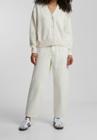 Osoba w off-white swetrze zapinanym na guziki, dopasowanych szerokich spodniach i białych trampkach z czarnymi paskami, stojąca na jednolitym szarym tle.