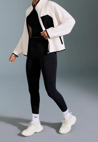 Osoba w czarnych legginsach, czarnym topie, białej kurtce z czarnymi detalami, białych skarpetkach Nike i białych sneakersach stoi na szarej podłodze.