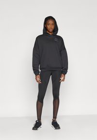 Crna majica s kapuljačom s logotipom na prsima, u kombinaciji s crnim tajicama koje imaju prozirni dizajn i bijele bočne pruge.