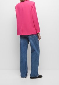 Personne portant un blazer rose vif, un jean bleu ample et des chaussures plates noires, debout de dos sur un fond blanc uni.