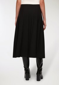 Gonna midi plissettata nera realizzata in tessuto leggero, con una vita liscia che si abbassa elegantemente fino alle caviglie. Indossata con stivali sopra il ginocchio.