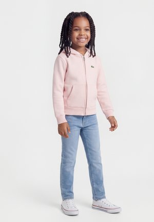 Ragazza sorridente con capelli intrecciati che indossa una felpa rosa con cerniera, jeans azzurri e sneakers bianche, in piedi davanti a uno sfondo semplice.