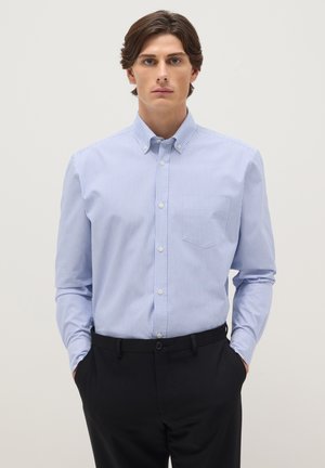 Man draagt een lichtblauw-wit gestreept overhemd met knopen en een zwarte broek, staand met handen in de zakken tegen een effen achtergrond.