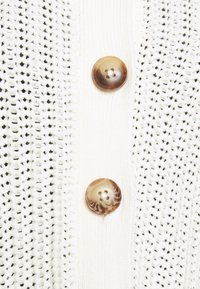 Vit stickad kofta med öppen struktur, försedd med två runda knappar med marmorerat mönster. Enkel design med en ren finish.