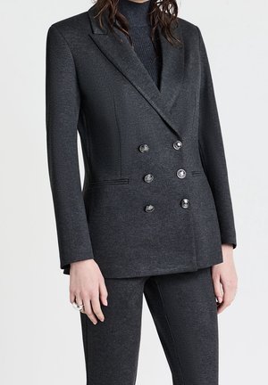 Kvinde iført en mørkegrå dobbeltradet blazer med seks knapper over matchende tætsiddende bukser og en sort rullekravebluse.